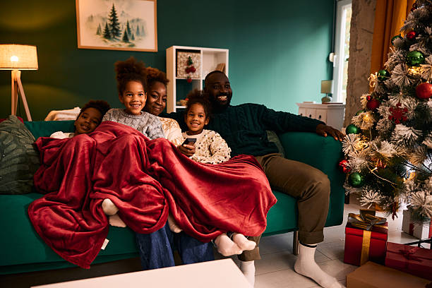 Loving African American family of five, wrapped in a cozy red blanket, enjoying Christmas holidays together while watching television on the sofa in their decorated living room